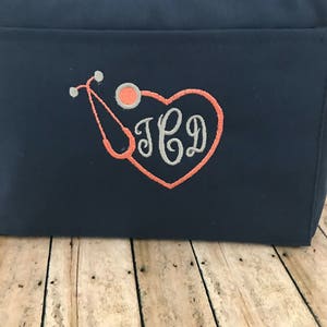 May include: A navy blue tote bag with a coral heart and stethoscope design. The bag is embroidered with the initials "JCD".