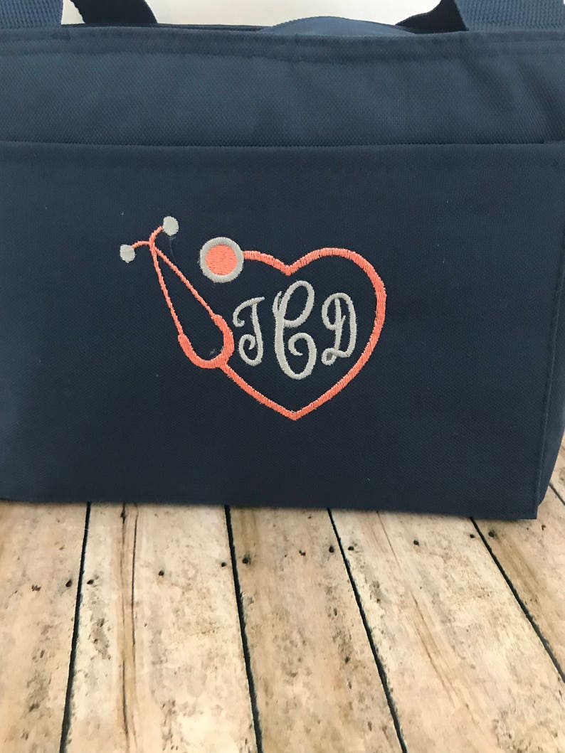 May include: A navy blue tote bag with a coral heart and stethoscope design. The bag is embroidered with the initials "JCD".