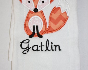 Puede incluir: Manta de bebé blanca con un diseño de zorro naranja bordado. El zorro tiene un patrón de chevrón y el nombre "Gatlin" está bordado en escritura negra.