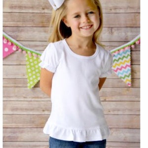 May include: A white short-sleeved t-shirt with a ruffled hem. The shirt is made of a soft, comfortable material and has a round neckline. The girl is wearing blue jeans and a white bow in her hair.