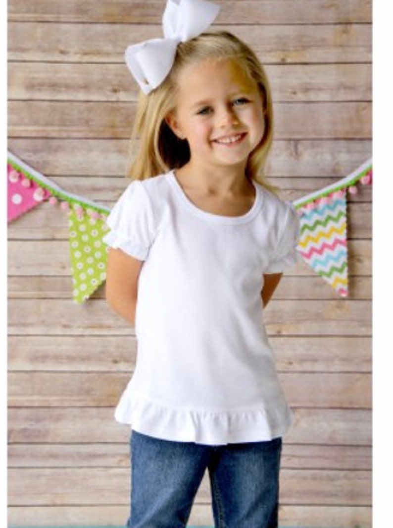 May include: A white short-sleeved t-shirt with a ruffled hem. The shirt is made of a soft, comfortable material and has a round neckline. The girl is wearing blue jeans and a white bow in her hair.