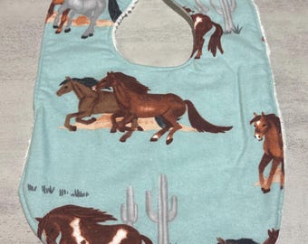 Puede incluir: Un babero para bebé con un fondo azul claro con caballos marrones y grises y cactus. El babero tiene un borde blanco y una forma curva diseñada para atrapar derrames. Los caballos están en varias poses, algunos corriendo y otros de pie.