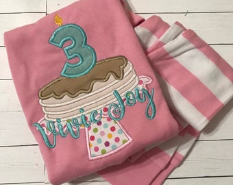 Puede incluir: Camiseta rosa de manga larga con una tela a rayas blancas y rosas debajo. La camiseta tiene un diseño bordado de un pastel con una vela número 3 y el texto "Vivie Joy".