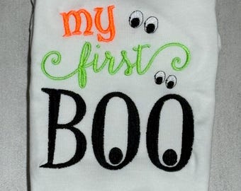 May include: A white garment featuring embroidered text that reads my first BOO. The word my is in orange, first is in lime green cursive, and BOO is in large black letters. Two pairs of cartoon eyes are embroidered above the word first and BOO.