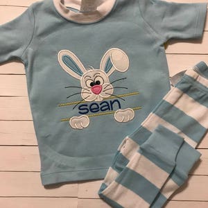 May include: Light blue pajama set featuring a bunny design with the name "Sean" embroidered below. The short-sleeved top has a white collar and the matching pants have blue and white stripes. The bunny has a pink nose and blue ears.