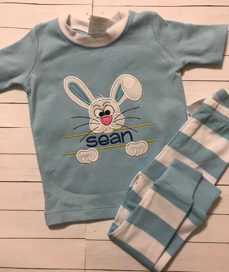 May include: Light blue pajama set featuring a bunny design with the name "Sean" embroidered below. The short-sleeved top has a white collar and the matching pants have blue and white stripes. The bunny has a pink nose and blue ears.