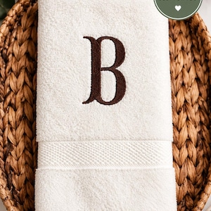 May include: A white cotton hand towel featuring a dark brown embroidered letter B in a serif font. The towel has a textured border near the bottom edge and is folded neatly. A circular green badge with the text PERSONALIZED JUST FOR YOU is visible.