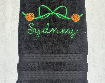 Puede incluir: Una toalla de mano negra con el texto bordado en verde que dice Sydney, situado bajo un lazo bordado en verde flanqueado por dos balones de baloncesto naranjas. La toalla tiene una superficie de rizo texturizado y un borde acanalado horizontal.
