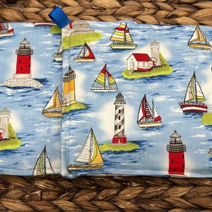 May include: A pair of blue and white cotton pot holders with a nautical theme. The pot holders feature lighthouses, sailboats, and islands. They have blue ribbon loops for hanging.