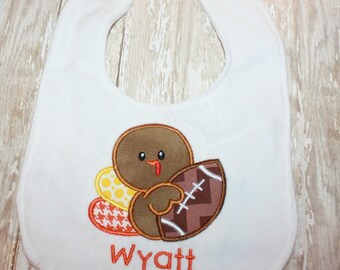 Puede incluir: Un babero blanco para bebé con un pavo marrón bordado que sostiene un balón de fútbol americano. El pavo tiene plumas de cola amarillas con lunares y naranjas con estampado de pata de gallo. El nombre Wyatt está bordado en naranja.