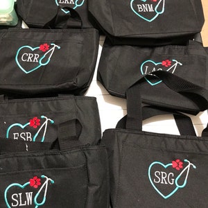 May include: Black tote bags with embroidered designs. Each bag features a heart with initials, a paw print, and a stethoscope. The bags have black handles and a front pocket. The embroidery is in shades of blue, red, and white.