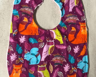 May include: A purple baby bib featuring a repeating pattern of orange foxes, light blue squirrels, and owls in shades of pink and green, accented with colorful leaves and acorns. The bib includes a purple snap closure at the top neck opening.