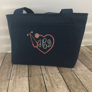 May include: Navy blue tote bag with a front pocket featuring an embroidered stethoscope and heart with the monogram "JCD".