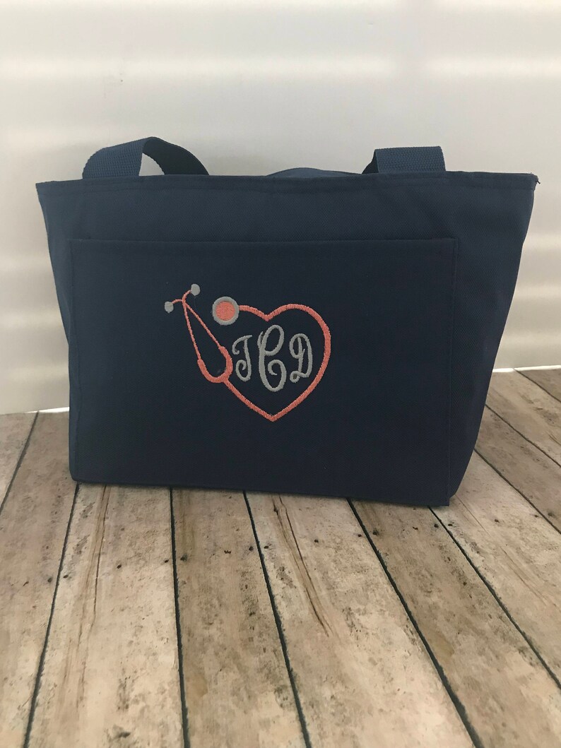 May include: Navy blue tote bag with a front pocket featuring an embroidered stethoscope and heart with the monogram "JCD".