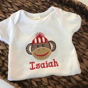 May include: White baby onesie with a red and brown embroidered monkey wearing a red and white striped hat. The onesie is personalized with the name "Isaiah" in red embroidery.