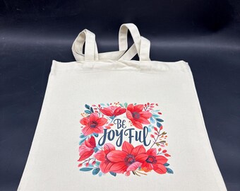 Puede incluir: Una bolsa de tela color crema con dos asas de tela y un estampado gráfico central. El diseño muestra flores rojas y rosas con hojas verdes que rodean el texto azul oscuro que dice Be Joy Ful en una fuente cursiva estilizada.