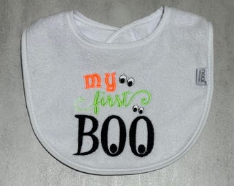 May include: A white baby bib featuring embroidered text that reads my first BOO in orange, light green, and black thread. The design includes two pairs of small cartoon eyes integrated into the lettering. A small grey tag is attached to the side.