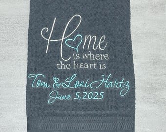 Puede incluir: Una toalla de mano gris oscuro con bordados en blanco y azul claro. El texto dice "Home is where the heart is" con un icono de corazón, seguido de los nombres "Tom & Loni Hartz" y la fecha "June 5, 2025" en una fuente cursiva.