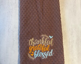 May include: A brown waffle-weave kitchen towel featuring embroidered text that reads "thankful grateful blessed" in gold, orange, and light blue thread, accented with small embroidered leaf and heart motifs above and below the words.