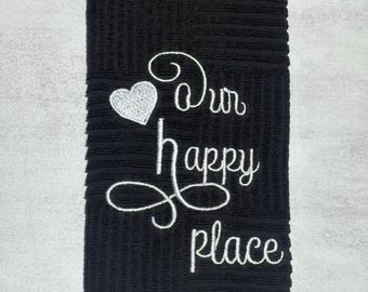 May include: A black rectangular kitchen towel with a textured ribbed surface. The towel features white embroidered text that reads Our happy place, accompanied by a small white embroidered heart icon positioned to the left of the word Our.