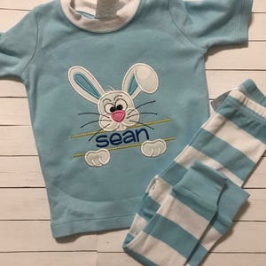 May include: Light blue children's pajama set featuring a cartoon bunny face with the name "sean" embroidered below. The top has a white collar and short sleeves. The pajama pants are light blue and white striped.