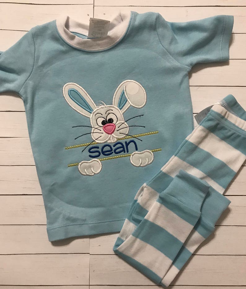 May include: Light blue children's pajama set featuring a cartoon bunny face with the name "sean" embroidered below. The top has a white collar and short sleeves. The pajama pants are light blue and white striped.