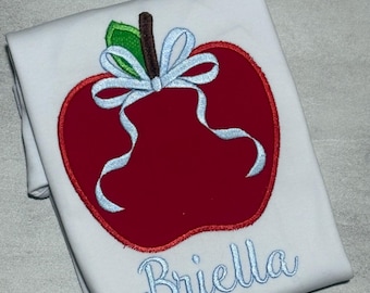 May include: A white t-shirt featuring an embroidered red apple with a brown stem and a green leaf. A light blue ribbon is tied around the stem. The name Briella is embroidered in light blue cursive script below the apple design.