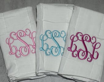 May include: Three white rectangular fabric cloths, each featuring a centered embroidered monogram in a cursive script. The left cloth has a light pink monogram, the center cloth has a light blue monogram, and the right cloth has a dark pink monogram.