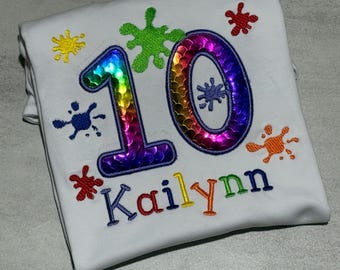 May include: A white t-shirt featuring an embroidered number 10 in iridescent rainbow sequins. The design includes colorful paint splatters in yellow, green, red, blue, and orange, with the name Kailynn embroidered in multicolored letters below.