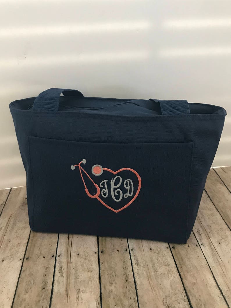 May include: A navy blue tote bag with a zipper closure and a large pocket on the front. The pocket features an embroidered design of a stethoscope and a heart with the monogram "J.C.D."
