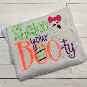 May include: A light gray Halloween-themed shirt with the phrase "Shake your BOOty" embroidered in green, orange, and purple. A white ghost with a pink bow is also embroidered on the shirt. The shirt is folded on a light wood surface.