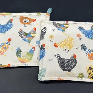 May include: Two white cotton pot holders with a colorful chicken pattern. The chickens are in various poses and have different patterns on their feathers. The pot holders have a blue and white trim.