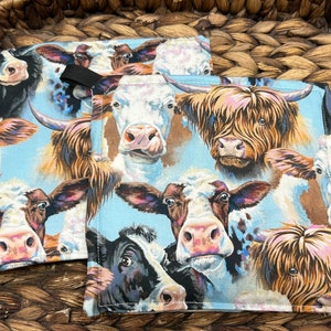 May include: Two square fabric pot holders with a cow-themed pattern. The design features various cow faces in shades of brown, black, and white against a light blue background. Each pot holder has a black loop for hanging.