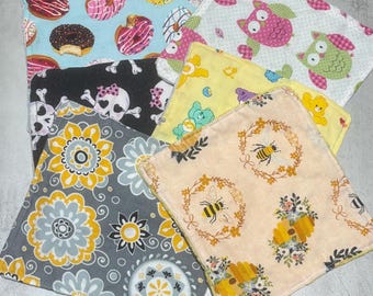 Puede incluir: Una colección de seis paños de tela cuadrados con varios patrones, incluyendo donas coloridas sobre azul claro, búhos rosas y verdes sobre blanco, calaveras negras, osos de dibujos animados amarillos, medallones florales grises y abejas sobre fondo melocotón.