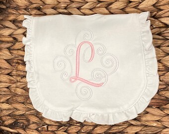 Puede incluir: Un babero de tela blanca con un borde con volantes y un diseño bordado central. El bordado consiste en una letra L grande de color rosa rodeada de patrones decorativos blancos en forma de espiral cosidos en el centro de la tela.