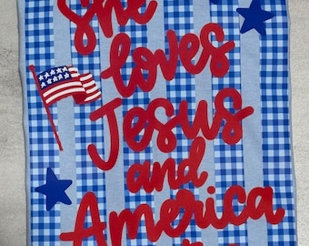 Puede incluir: Un paño de cocina a cuadros azul claro y blanco con el texto "She loves Jesus and America too" en escritura roja. El paño de cocina también presenta una pequeña bandera estadounidense y estrellas azules.