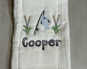May include: A white burp cloth featuring an embroidered design of a fishing rod with a light blue fish, flanked by two green cattails. Below the embroidery, the name Cooper is stitched in dark blue, bold, rounded, lowercase lettering.