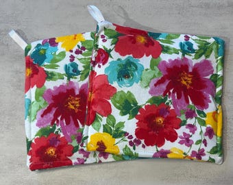 Puede incluir: Dos agarradores de cocina cuadrados y acolchados con un vibrante estampado floral sobre fondo blanco. El diseño incluye flores grandes de color rojo, magenta, amarillo y turquesa con hojas verdes. Cada agarrador tiene una pequeña trabilla blanca.