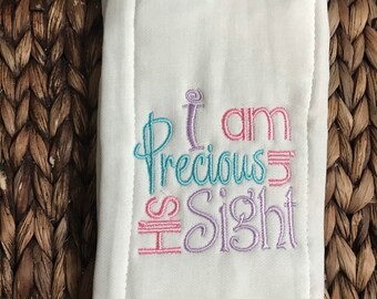 Puede incluir: Un paño de eructos rectangular blanco con texto bordado que dice I am Precious in His Sight. El bordado utiliza una mezcla de hilo azul claro, morado y rosa en una fuente cursiva decorativa, centrada en la superficie de la tela.
