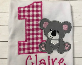 Könnte beinhalten: Weißer Strampler mit einer pink-weißen Gingham-Zahl eins und einem grauen Koalabären. Der Name "Claire" ist in Pink unter dem Koala gestickt.