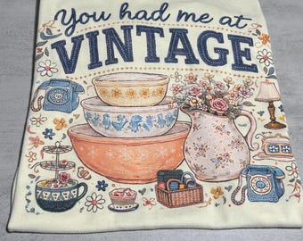 Puede incluir: Una camiseta de color crema con el texto "You had me at VINTAGE" en azul oscuro. El gráfico incluye ilustraciones de cuencos apilados, una jarra floral, teléfonos de disco, una lámpara, una cesta y varias flores pequeñas.