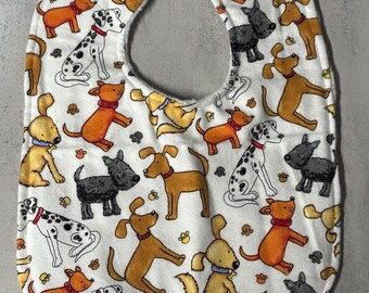 May include: A white baby bib featuring a repeating pattern of various dog breeds in shades of orange, black, and white. The illustrated dogs are depicted in different poses, including sitting and standing, with small paw prints scattered throughout.