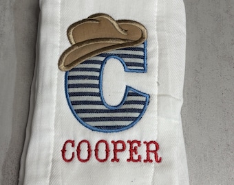 May include: A white burp cloth featuring an embroidered design of a tan cowboy hat resting on top of a large letter C with blue and white horizontal stripes. Below the letter C, the name COOPER is embroidered in red capital letters.
