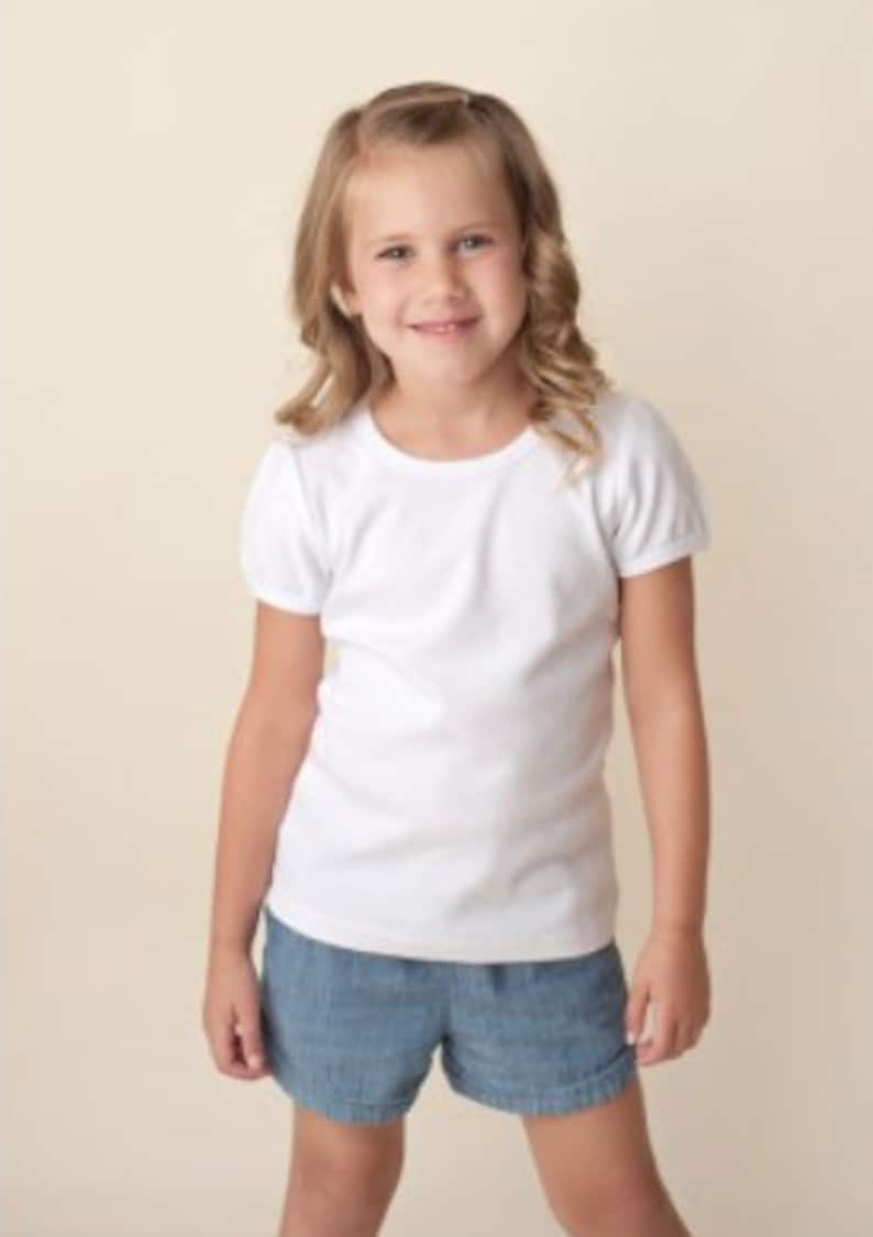 May include: A young girl wearing a white short-sleeved t-shirt and blue denim shorts.