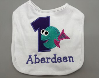 Puede incluir: Babero blanco para bebé con el número 1 morado y un pez turquesa con aletas rosas. El nombre "Aberdeen" está bordado en morado debajo. Un babero colorido para el primer cumpleaños de un bebé.