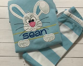 Puede incluir: Un conjunto de pijama azul claro con una cara de conejo de dibujos animados y el nombre "sean" bordado en azul. El conejo tiene orejas blancas con detalles azules, una nariz rosa y bigotes negros. El conjunto incluye pantalones a rayas azules y blancas.