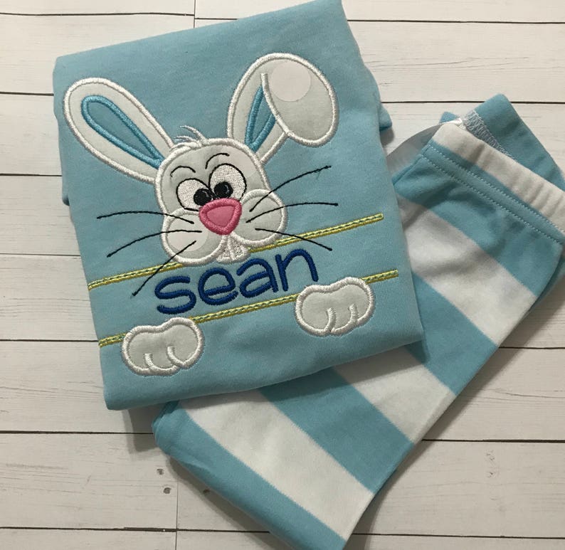 May include: A light blue pajama set featuring a cartoon bunny face with the name "sean" embroidered in blue. The bunny has white ears with blue accents, a pink nose, and black whiskers. The set includes striped blue and white pants.