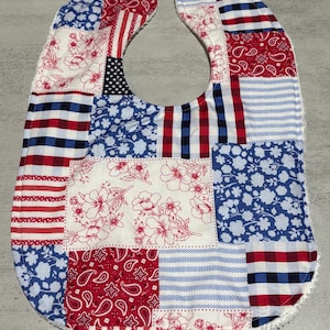 May include: A patchwork baby bib with a patriotic theme. The bib features various fabric patterns, including red and white stripes, blue floral prints, and a section with the American flag. The bib is edged with white terry cloth and has a snap closure.
