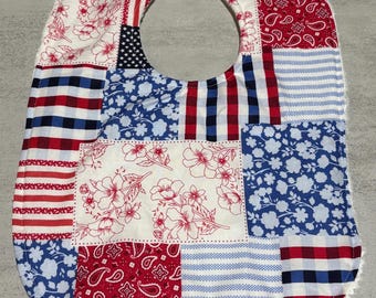 Puede incluir: Babero de bebé patchwork con temática patriótica. El babero presenta varios patrones de tela, incluyendo rayas rojas y blancas, estampados florales azules y una sección con la bandera americana. Tiene un borde de rizo blanco y un cierre de botón a presión.
