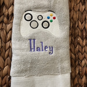 May include: A light gray hand towel with a white embroidered video game controller design. The controller has black outlines and colorful buttons. The name "Haley" is embroidered in purple below the controller.